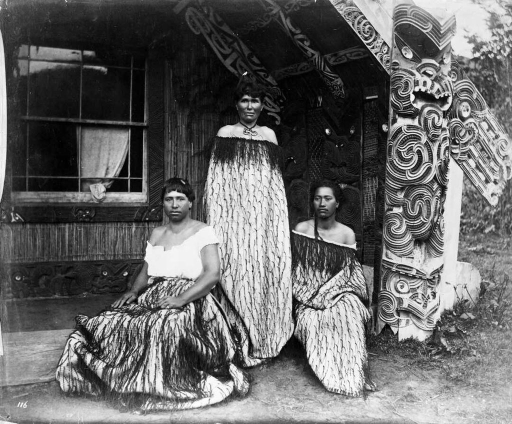 Tourist guides Sophia Hinerangi (standing), Kate Middlemass (Kati) (on left) and another guide, outside Hinemihi meeting house at Te Wairoa. Taken by Elizabeth Pulman around 1881.